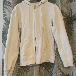 Vit hoodie med dragkedja och huva - Snygg vit hoodie med dragkedja framtill och huva med snörning. Tröjan har två stora fickor på magen och långa ärmar. Perfekt för en chill och avslappnad stil.