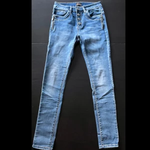 Blå skinny jeans från Chica London - Snygga blå skinny jeans från Chica London med hög midja och flera knappar framtill. Jeansen har coola dragkedjor vid fickorna och en liten stjärndetalj på bakfickan. Perfekt passform och stretchigt denimtyg som sitter tajt hela vägen ner.