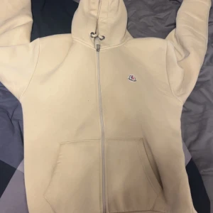 Beige hoodie från Moncler med dragkedja - Säljer en beige hoodie från Moncler med hel dragkedja, huva med snörning och två stora fickor framtill. Tröjan har ett litet Moncler-märke på bröstet och är tillverkad i mjuk bomull. Perfekt för chill dagar och snygg till jeans.