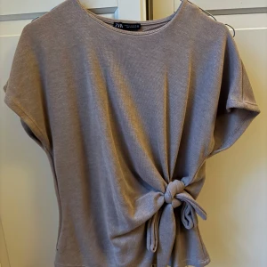 Beige topp med knyt från Zara - Säljer en beige topp från Zara med korta ärmar och rund hals. Toppen har en snygg knytning i sidan som ger en cool och avslappnad look. Perfekt till jeans eller kjol för en trendig vardagsstil.