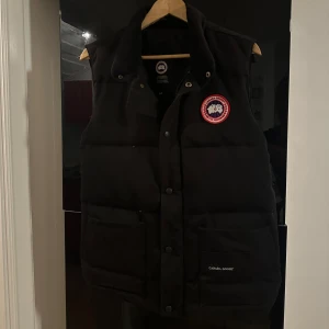 Svart dunväst från Canada Goose - Svart dunväst från Canada Goose med klassisk logga på bröstet i storlek S, västen har inte använts så mycket men säljs nu då den ej passar min stil längre. 