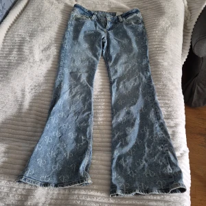 Blåa bootcut jeans från H&M Divided - Snygga ljusblå bootcut jeans från H&M Divided med all-over tryckt mönster. Jeansen har klassiska fickor fram och bak, samt coola detaljer med knappar på bakfickorna. Perfekta för dig som vill sticka ut med en unik denimstil.