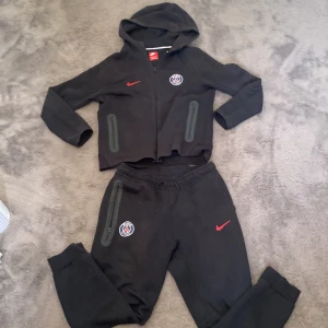 Svart PSG hoodie och mjukis byxor från Nike  - Svart hoodie från Nike med PSG-logga på bröstet och röd swoosh. Tröjan har huva, dragkedja framtill och två fickor med zip. Perfekt för dig som gillar fotboll och vill ha en sportig look. Materialet är mjukt och känns skönt mot huden.