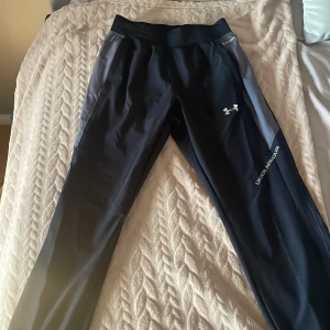 Svarta träningsbyxor från Under Armour - OANVÄNDA, har aldrig använt byxorna. Snygga svarta träningsbyxor från Under Armour med grå detaljer på sidorna och logga på låret. Byxorna har elastisk midja och är gjorda i ett lätt, syntetiskt material som andas. Perfekta för dig som vill ha bekväma och funktionella byxor till träningen.