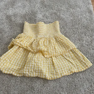 Gul rutig volangkjol från KappAhl - Supersöt kort kjol från KappAhl i gul och vit rutig bomull. Kjolen har bred smock i midjan och dubbla volanger som ger en riktigt härlig sommarkänsla. Perfekt för dig som gillar färg och mönster!