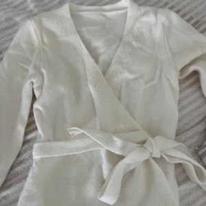 Vit omlottkofta med knytband - Mysig vit omlottkofta med knytband i midjan och långa ärmar. Koftan är stickad och har en v-ringad design som ger en clean och stilren look. Perfekt att slänga på sig över en topp för en chill och snygg outfit. Pris kan diskuteras 