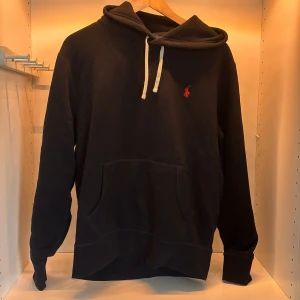 Svart hoodie från Ralph Lauren - Svart hoodie från Ralph Lauren med klassisk röd logga broderad på bröstet. Tröjan har huva med vita snören och en stor magficka framtill. Långärmad och i mjuk bomull, perfekt för en chill och stilren look. Ny pris 2000kr