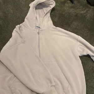 Ljuslila hoodie från Kappahl - Ljuslila hoodie från Kappahl med dragkedja framtill och stor huva. Tröjan har en klassisk känguruficka och ribbade muddar vid ärmslut och nederkant. Perfekt för en chill och avslappnad stil.