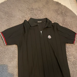 Svart t-shirt med dragkedja - Svart t-shirt med korta ärmar, diskret lodrätt ribbad struktur och klassisk krage. Dragkedja framtill och broderad på bröstet. Ärmsluten har snygga rödvita detaljer som ger en sportig vibe.