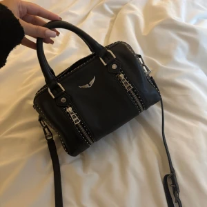 Svart handväska Zadig & Voltaire skinn - Cool svart handväska i skinn från Zadig & Voltaire med silverfärgade detaljer, dubbla dragkedjor framtill och nitar längs kanterna. Axelremmen har broderad logga och väskan har ett stilrent emblem med vingar på framsidan. Perfekt storlek för vardagens essentials.