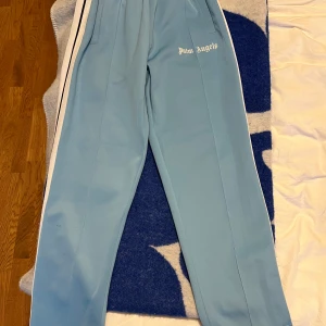 Palm Angels ljusblå trackpants - Snygga ljusblå trackpants från Palm Angels med vita sidoränder och logga tryckt på låret. Byxorna har elastisk midja och raka ben, tillverkade i ett glansigt polyestermaterial som ger en sportig vibe. Perfekta för en streetwear-look.