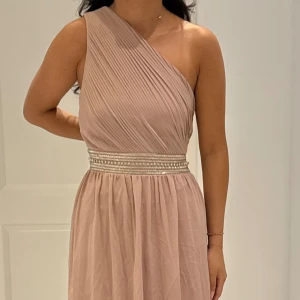 Dusty rosé långklänning med pärldetaljer - Elegant dusty rosé långklänning med plisserad överdel och bred midja dekorerad med pärlor och glittriga detaljer. Klänningen har en snygg oneshoulder-skärning och faller mjukt i ett tunt, svepande tyg. Perfekt för bal eller festligheter. Helt ny med prislappen på!