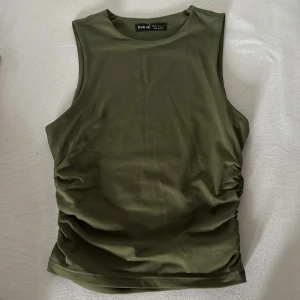 Topp - Snygg olivgrön ärmlös topp med rund halsringning och rynkade sidor som ger en cool och modern look. Tillverkad i mjukt och stretchigt syntetmaterial, perfekt för att lyfta din outfit med en enkel men trendig vibe.