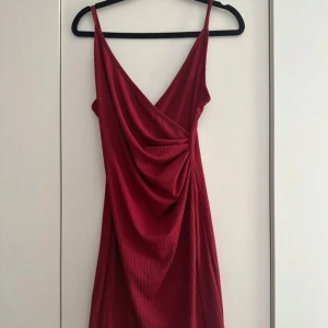 Vinröd omlottklänning med smala axelband - Vinröd klänning med omlott och draperad front, smala justerbara axelband och subtilt ribbat mönster. Klänningen har v-ringning och är tillverkad i ett mjukt, stretchigt material som ger en snygg passform
