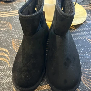 Svarta mockaboots med platt sula - Svarta boots i mocka med rund tå och platt sula. Skorna har en enkel och stilren design med kort skaft och mjukt foder på insidan. Perfekta för kalla dagar när du vill ha både värme och stil.