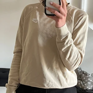 Beige sweatshirt från Nike - Säljer en stilren beige sweatshirt från Nike med broderad vit logga på bröstet. Tröjan har rund halsringning och långa ärmar, perfekt för en avslappnad och sportig look. Använd fåtal gånger, nypris 549kr