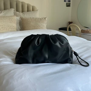 Svart clutch i skinnimitation - Säljer en svart clutch med mjuk, puffig form och snyggt rynkad design. Väskan stängs med en dragkedja och har ett innerfack. Passar till vardags och fest!!🎉🩷
