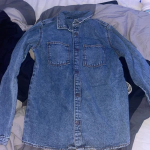 Jeansskjorta från Primark, 152 cm - Snygg jeansskjorta från Primark i klassisk blå denim. Skjortan har två bröstfickor, knappar framtill och långa ärmar. Perfekt för en chill och avslappnad look. Passar dig som gillar enkel och tidlös stil.