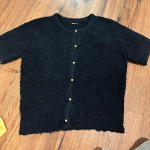 Topp från Gina Tricot - Mysig mörkblå kofta från Gina Tricot med korta ärmar och dekorativa knappar framtill. Koftan har en fluffig, stickad känsla och rund halsringning. Perfekt för lager-på-lager och enkel att styla till olika looks.