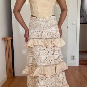 Beige ruffles embroidered skirt - Superfin beige långkjol i 100% bomull med broderade detaljer och volanger. Kjolen har vackert hålbroderi, spetsinslag och flera lager volanger som ger en romantisk vibe. Perfekt för dig som gillar boho-stil och vill sticka ut med unika detaljer.