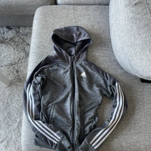 Grå Adidas kofta med huva XS - Snygg grå kofta från Adidas i storlek XS. Koftan har huva, hel dragkedja och klassiska vita ränder längs ärmarna. Perfekt för träning eller chill dagar. Adidas-logga på bröstet och två fickor framtill.