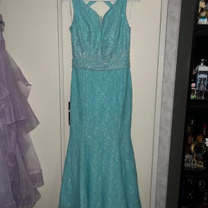 Turkos långklänning med pärlor och spets - Säljer en turkos långklänning med spets och glittriga pärlor upptill. Klänningen har v-ringning, korsade axelband i ryggen och dragkedja bak. Kjolen är i spets och har en elegant, figurnära passform. Perfekt för bal eller festligheter! Gick lite sönder i mitten men inget som syns jättemycket och det går att fixa! Storlekslappen säger 40 men jag hade sagt att den passar mer en 38💗