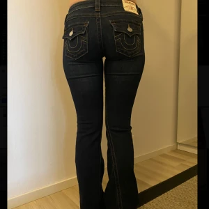 Mörkblå True Religion bootcut jeans - Snygga mörkblå jeans från True Religion med bootcut passform och kontrastsömmar. Byxorna har låg midja och fickdetaljer bak med lock och knappar. Säljes då de är lite för långa för mig som är 166cm💕Jag brukar vanligtvis ha XS/S i jeans. Nypris 1300kr