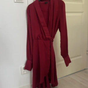 Vinröd omlottklänning Vero Moda S - Supersnygg vinröd omlottklänning från Vero Moda i storlek S. Klänningen har långa ärmar, knyt i midjan och v-ringad hals. Den är kort och har en flowy passform som ger en chic vibe. Perfekt för dig som vill sticka ut med en djup röd färg och stilren look. Passar även super vid olika tillfällen som julafton