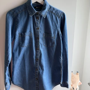 Jeansskjorta från Gina Tricot, stl 36 - Snygg jeansskjorta från Gina Tricot i klassisk blå denim. Skjortan har två bröstfickor, långa ärmar med knappar vid manschetterna och genomgående knäppning framtill. Perfekt till ett par svarta jeans eller kjol för en cool look.
