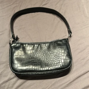 Svart axelväska i croco från H&M Divided - Snygg svart axelväska från H&M Divided med croco-mönstrad yta och silvrig dragkedja. Väskan har en stilren, avlång form och bred axelrem. Perfekt för att lyfta din outfit med en trendig touch.