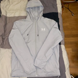 Under Armour Rival jacka (ny) - Snygg ljusgrå vindjacka från Under Armour med huva och dragsko. Jackan har dragkedja framtill, två sidofickor och meshfoder för extra komfort. Perfekt för dig som gillar sportig stil och vill ha något lätt och smidigt perfekt till sommaren.