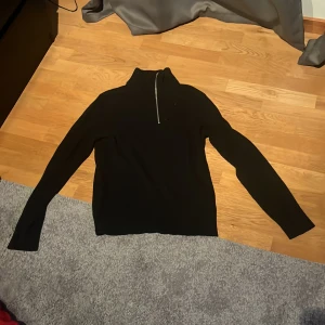 Svart stickad tröja med half zip - Svart stickad tröja i storlek S med hög krage och snygg half zip-dragkedja. Tröjan har lång ärm och en clean, minimalistisk look som passar perfekt till jeans eller kjol. Materialet är mjukt och ger en skön känsla hela dagen.