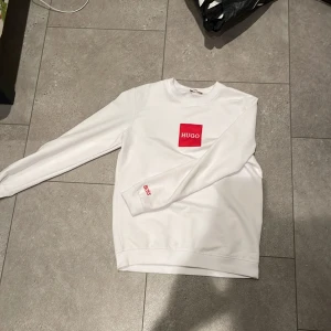 Vit sweatshirt från Hugo Boss - Snygg vit sweatshirt från Hugo Boss med röd HUGO-logga på bröstet och BOSS-detalj på ärmen. Tröjan har rund halsringning, långa ärmar och är tillverkad i mjuk bomull. Perfekt för en clean och stilren look.