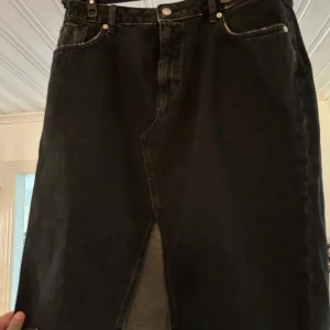 Svart jeanskjol med slits - Svart jeanskjol med rå kant och cool slits framtill. Klassisk femficksmodell med bälteshällor och bakfickor. Perfekt för dig som gillar en edgy och avslappnad stil. Storlek L