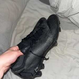 Svarta Nike Mercurial fotbollsskor - Säljer ett par helsvarta Nike Mercurial fotbollsskor med snörning och platt sula med dobbar. Skorna har en strömlinjeformad design och är tillverkade i syntetmaterial med textilinslag för en lätt känsla på planen.