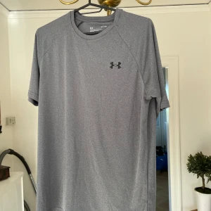 tränings-tshirt Under Armour - Säljer en grå t-shirt från Under Armour i loose fit. Perfekt för träning med sitt lätta och snabbtorkande polyestermaterial. Klassisk rund hals, kort ärm och diskret logga på bröstet och nacken. Enkel och stilren design som funkar till alla sportiga outfits. Stlk S. Använd 1 gång!
