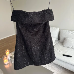 Svart axelbandslös kort klänning H&M - Snygg svart axelbandslös kort klänning från H&M med glittrig struktur och markerad krage upptill. Klänningen har dragkedja bak och är fodrad för extra komfort. Perfekt för dig som vill sticka ut med en elegant och trendig look.