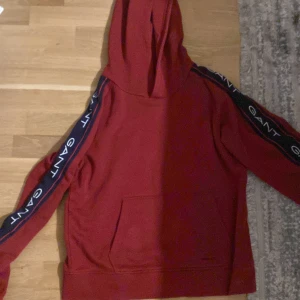 Röd hoodie från GANT med logga - Röd hoodie från GANT med huva och känguruficka. Snygga marinblå ränder med GANT-logga längs båda ärmarna ger en sportig vibe. Perfekt för chill dagar eller när du vill sticka ut lite extra.