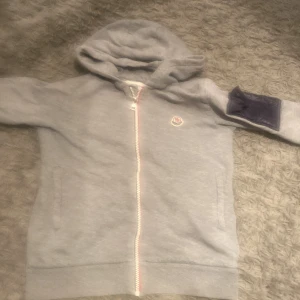 Grå Moncler hoodie med dragkedja - Grå hoodie från Moncler med hel dragkedja och huva. Klassisk logga på bröstet och en mörkblå patch på ena ärmen. Mjuk bomullskänsla och två fickor framtill. Perfekt för chill dagar och streetstyle.