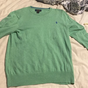 Grön tröja från Polo Ralph Lauren - Snygg grön tröja från Polo Ralph Lauren med klassisk blå logga broderad på bröstet. Tröjan har rund halsringning, långa ärmar och ribbade muddar vid ärmslut och nederkant. Perfekt för dig som gillar stilren och enkel design.