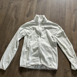 Vit fleecejacka med dragkedja - Mysig vit fleecejacka med hel dragkedja och hög krage. Perfekt för kyliga dagar och enkel att slänga på sig över en hoodie eller t-shirt. Jackan har långa ärmar och två sidofickor. Stilren och enkel design som funkar till det mesta.