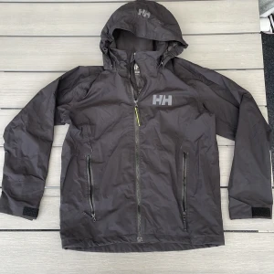 Svart vindjacka/regnjacka från Helly Hansen - Svart vindjacka från Helly Hansen med huva, hel dragkedja och två sidofickor med dragkedjor. Jackan har HH-logga på bröstet och huvan samt justerbara ärmslut med kardborre. Perfekt för blåsiga och regniga dagar och enkel att matcha med allt. Har även en luva som som går att ta av och stoppa in under kragen.