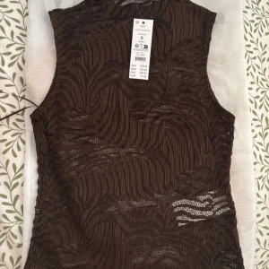 Brun ärmlös topp med mönster - Säljer en brun ärmlös topp med transparenta partier och vågigt mönster. Toppen har en tight passform och är tillverkad i ett syntetmaterial som ger en cool struktur. Perfekt för dig som vill sticka ut med en unik look.