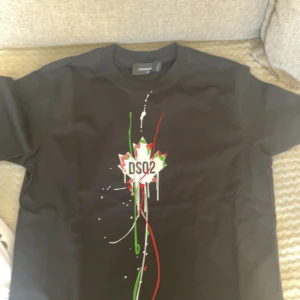 Svart DSQUARED2 t-shirt med print - Svart t-shirt från DSQUARED2 med coolt print framtill i form av ett lönnlöv och texten DSQ2. Trycket har detaljer i rött, grönt och vitt som ger en streetig vibe. Klassisk passform och rund hals. Perfekt för dig som gillar statement-plagg.