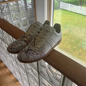 Maison Margiela sneakers med färgstänk - Unika vita sneakers från Maison Margiela med coola färgstänk i rött, blått, gult och svart. Skorna har snörning, platt sula i gummi och är tillverkade i skinn. Perfekt för dig som vill sticka ut med en kreativ och konstnärlig vibe.                                                   Priset är diskuterbart.