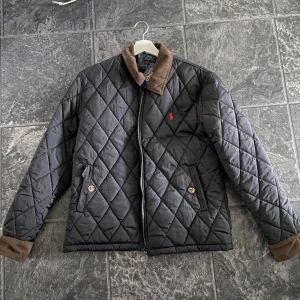 Svart quiltad jacka från Polo Ralph Lauren - Quiltad svart jacka från Polo Ralph Lauren med brunt manchesterdetaljer på krage och ärmslut. Klassisk röd logga på bröstet, två fickor med knappar och dragkedja framtill. Perfekt för kyliga dagar och snygg till jeans.