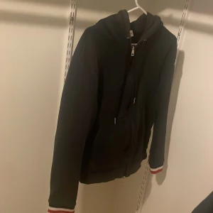 Mörkblå Moncler hoodie med dragkedja - Mörkblå hoodie från Moncler med hel dragkedja, huva och snörning. Röd och vit randig detalj på ärmslut och huva. Klassisk Moncler-logga på ena ärmen. Perfekt för dig som gillar stilrena och sportiga plagg.