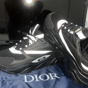 Svarta Dior sneakers med vita detaljer - Chunky svarta Dior sneakers med vita detaljer och logga på sidan. Ovandel i mesh och syntet med coola kontraster och kraftig sula. Skorna har snörning och en modern, sportig vibe som sticker ut. Perfekta för dig som gillar exklusiva streetwear-inspirerade skor.