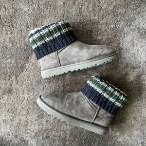 UGG x Sacai grå boots med stickad kant - Grå mockaboots från UGG x Sacai med ribbstickad kant i marinblått, grönt och vitt. Snyggt rutigt mönster upptill och platt sula. Perfekta för kalla dagar med sin mysiga och unika design. Strlk 42 passar 43/44