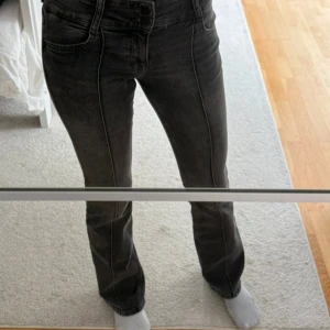 Grå bootcut jeans från Bershka - Säljer ett par gråa bootcut jeans från Bershka med låg midja och snygga söm-detaljer framtill. Jeansen har klassisk femficksdesign, dubbla knappar i midjan och är tillverkade i stretchigt denim för en bekväm passform.